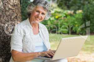 Cheerful mature woman using a laptop sitting on tree trunk