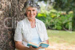 Cheerful mature woman holding book sitting on tree trunk