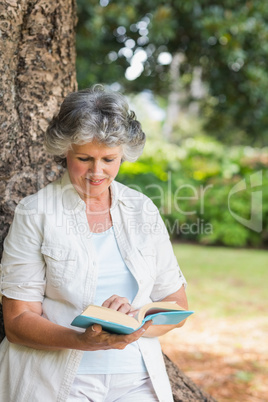 Cheerful mature woman reading book leaning on tree trunk