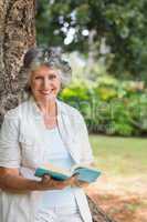 Smiling mature woman reading book leaning on tree trunk