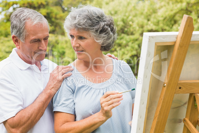Cheerful retired woman painting on canvas and talking with husba