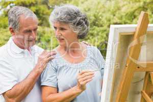 Cheerful retired woman painting on canvas and talking with husba
