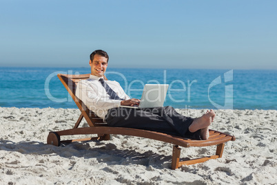 Smiling young businessman lying on a deck chair with his laptop