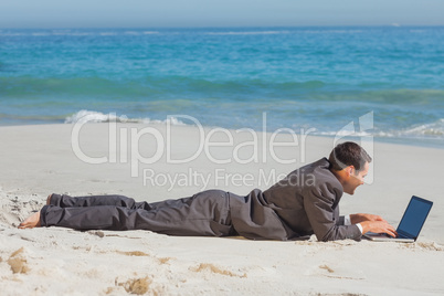 Young businessman lying on the sand typing on his laptop