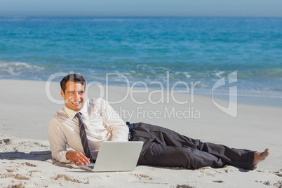 Smiling young businessman lying on the sand with his laptop
