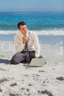 Pensive young businessman sitting on the sand with his laptop