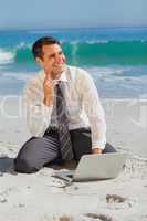 Thoughtful businessman sitting on the sand with his laptop