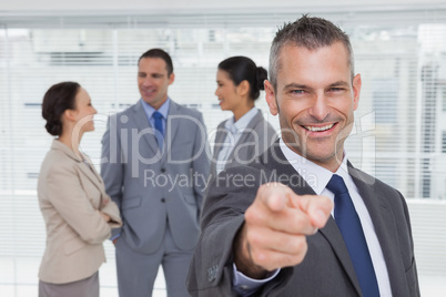 Cheerful businesman pointing at camera with colleagues on backgr