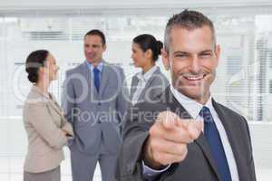 Cheerful businesman pointing at camera with colleagues on backgr