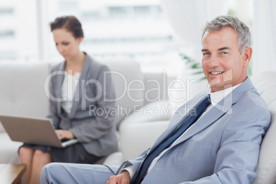 Businessman posing while his colleague working on her laptop