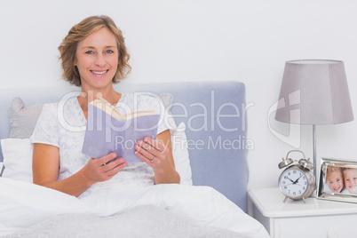 Relaxed blonde woman sitting in bed holding book