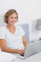 Smiling blonde woman sitting in bed using laptop