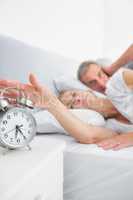 Tired wife turning off alarm clock as husband is covering ears