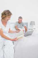 Happy woman reading book while husband is using laptop