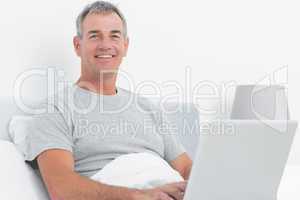 Happy grey haired man using his laptop in bed