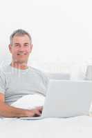 Cheerful grey haired man using his laptop in bed
