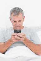 Smiling grey haired man sending a text in bed
