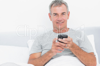 Cheerful grey haired man sending a text in bed