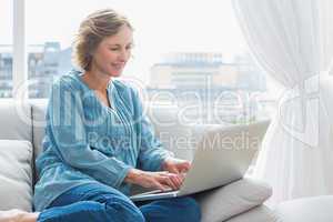 Happy blonde woman sitting on her couch using laptop