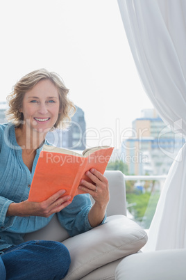 Happy blonde woman sitting on her couch holding a book