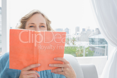 Blonde woman sitting on her couch covering face with orange book