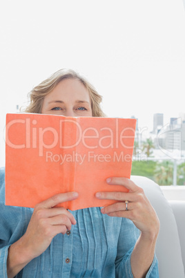 Woman sitting on her couch covering face with orange book