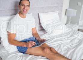 Smiling man using his tablet pc sitting on bed