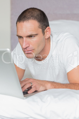 Serious man lying on bed using his laptop