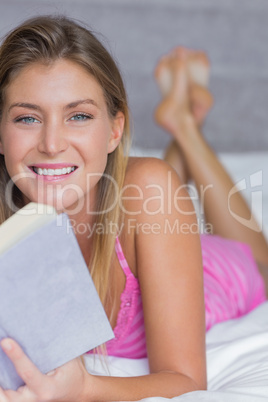Happy blonde lying on her bed reading a book smiling at camera