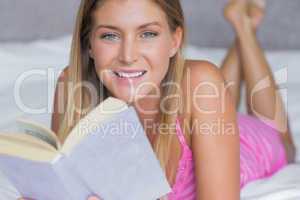 Pretty blonde lying on her bed reading a book smiling at camera