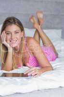 Happy blonde lying on her bed using tablet pc smiling at camera