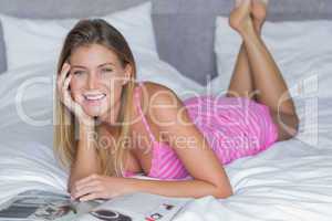 Beautiful blonde lying on her bed reading magazine smiling at ca