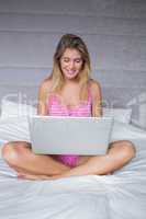 Happy blonde woman sitting on her bed using laptop