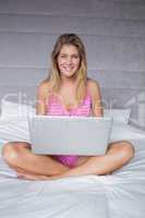Cheerful blonde woman sitting on her bed using laptop