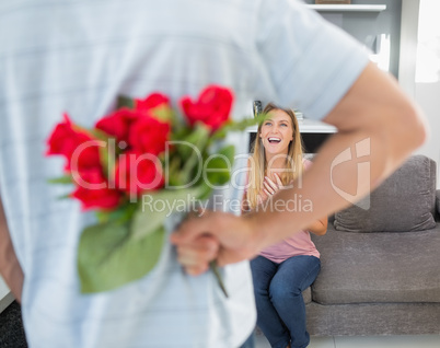 Man hiding bouquet of roses from smiling girlfriend on the couch