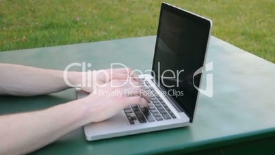 Hands type on a laptop that is resting on a green table outdoors