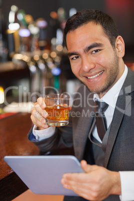 Cheerful businessman working on his tablet while having a whiske