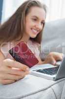 Smiling woman lying ona sofa holding a card and laptop