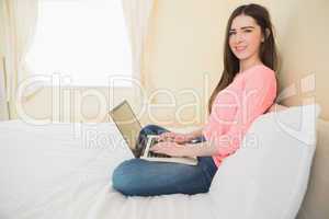 Happy girl looking at camera using a laptop sitting on a bed