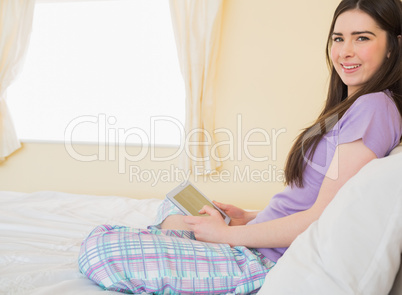 Relaxed girl sitting on a bed looking at camera and using a tabl