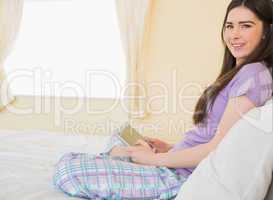 Relaxed girl sitting on a bed looking at camera and using a tabl