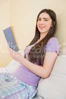 Cheerful girl looking at camera and sitting on a bed reading a b