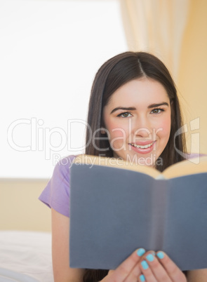 Happy girl looking at camera and lying on a bed reading a book