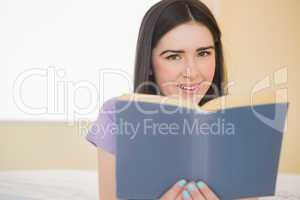 Cheerful girl looking at camera and lying on a bed reading a boo