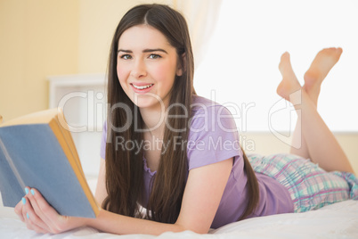 Pretty happy girl looking at camera and lying on a bed reading a