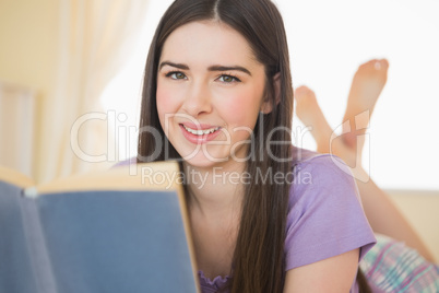 Pretty smiling girl looking at camera and lying on a bed reading