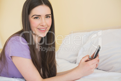 Cheerful girl looking at camera and lying on a bed using a mobil