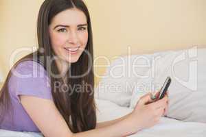 Cheerful girl looking at camera and lying on a bed using a mobil