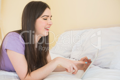 Cheerful brunette lying on a bed using a tablet pc in a bedroom