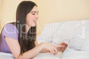 Cheerful brunette lying on a bed using a tablet pc in a bedroom